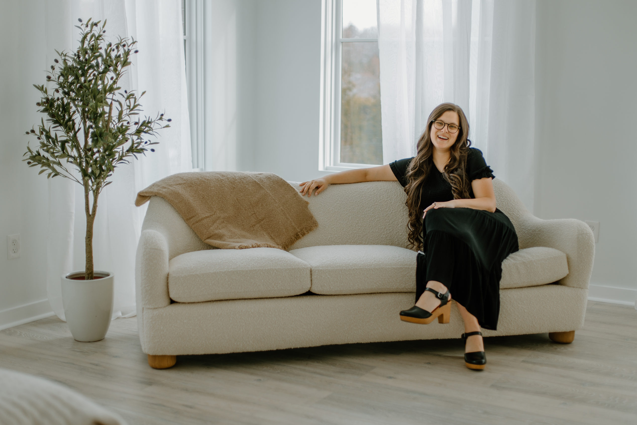 rachel, atlanta wedding photographer posing on a white couch next to a faux olive tree