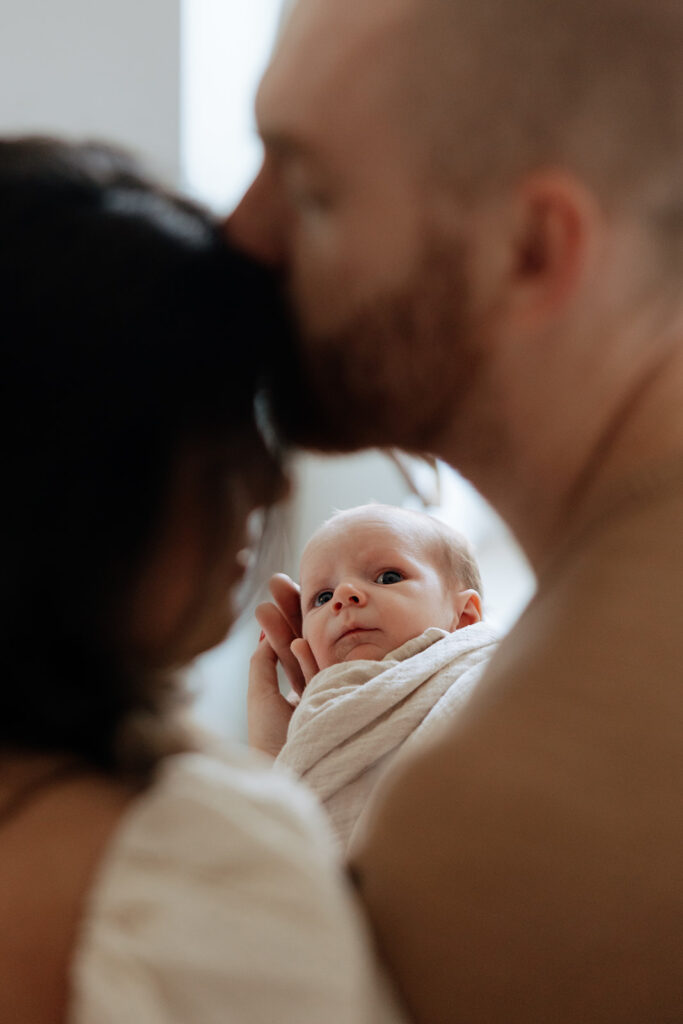 husband and wife kiss while holding new baby